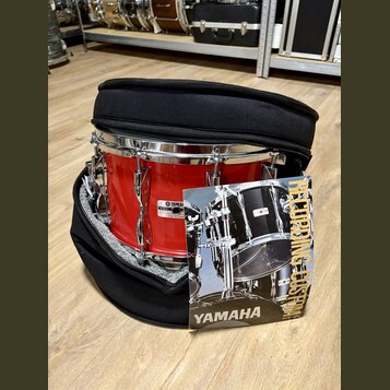 Červený buben v černém polstrovaném obalu. Na koruse je vidět štítek se značkou YAMAHA a nápisem MODEL: MADE IN JAPAN. Před nástrojem leží produktový…