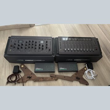 Dvě šedé studiové kontroléry Softube, model CONSOLE 1 CHANNEL Mk III a CONSOLE 1 FADER Mk III. Pod nimi jsou dvě krabice s nápisy Softube CONSOLE 1 C…