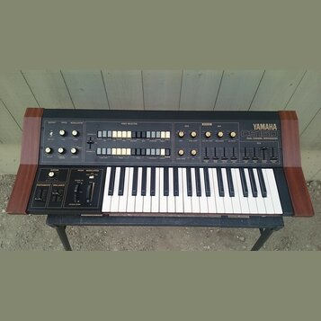 Černý analogový syntetizátor s dřevěnými bočními panely a bílo-černou klaviaturou. Na pravé straně je nápis YAMAHA CS15D DUAL CHANNEL SYNTHESIZER. Př…