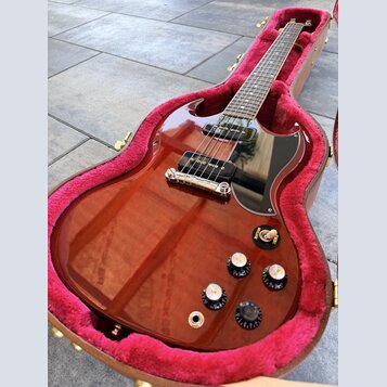 Elektrická kytara s červeně hnědým průsvitným lakem a černým pickguardem v otevřeném kufru s růžovou výstlkou. Nástroj má dva humbuckery, stříbrný ko…