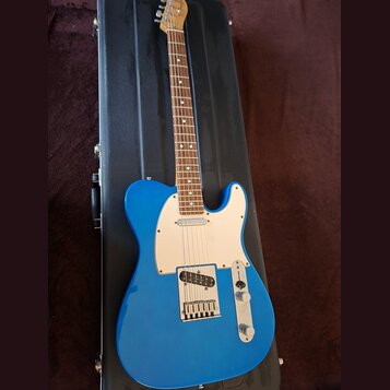Modrá elektrická kytara typu Telecaster s bílým pickguardem a hnedým hmatníkem, položená na černém tvrdém kufru. Nástroj má dva snímače a dva ovládac…
