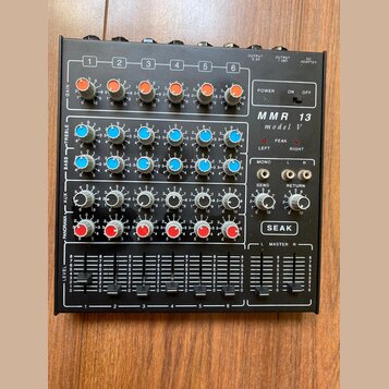 Černý mixážní pult s šesti kanály. Na pravé straně je bílý nápis MMR 13 model V a logo SEAK. Zařízení obsahuje řadu barevných otočných ovladačů v ora…