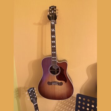 Akustická kytara s cutawayem v tmavě hnědém přechodovém odstínu. Na hlavě je nápis Gibson a na hmatníku jsou bílé vložky ve tvaru obdélníků. V místě…