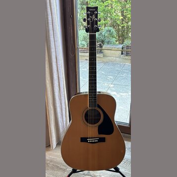 Akustická kytara s přirozenou dřevěnou barvou korpusu a černým hmatníkem. Na hlavě je bílý nápis YAMAHA a dekorativní ornament. Kytara má černý trsac…