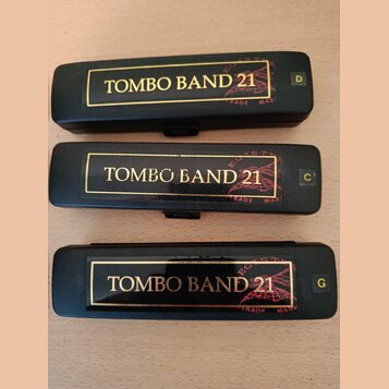 Tři černé harmoniky s nápisem TOMBO BAND 21 v zlatém rámečku. Každý nástroj má v pravém horním rohu označení tóniny: D, C a G. Na tělech jsou vidět n…