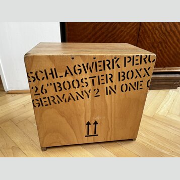 Dřevěný nástroj ve tvaru kostky s přirozenou barvou dřeva. Na přední straně je černý nápis SCHLAGWERK PERC 26"BOOSTER BOXX GERMANY2 IN ONE O a ve spo…