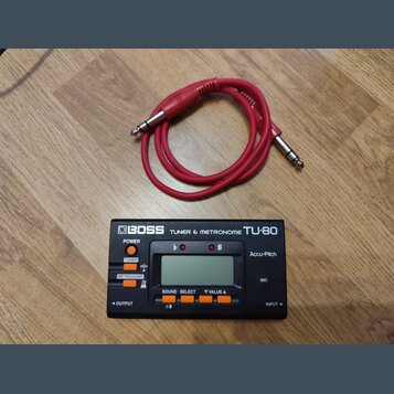 Černý digitální ladič a metronom značky BOSS, model TU-80. Zařízení má LCD displej, oranžová tlačítka a nápisy POWER, TUNER, METRONOME, SOUND SELECT,…