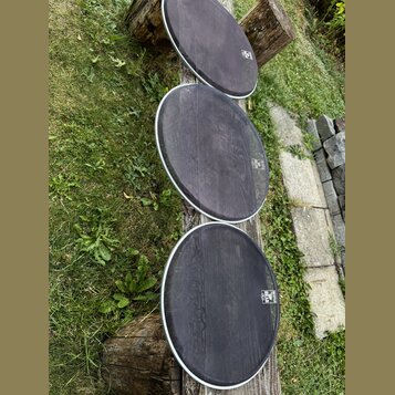 Tři kulaté bubnové membrány s bílým okrajem, položené na dřevěném trámku v trávě. Membrány jsou tmavé, s viditelnou strukturou povrchu a mírným opotř…