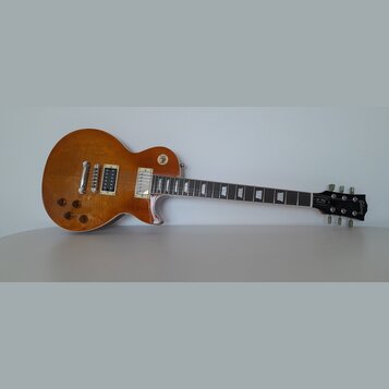 Elektrická kytara typu Les Paul v jantarové barvě s viditnou strukturou dřeva. Na hmatníku jsou trapezoidové vložky a na hlavě je nápis Gibson. Kytar…
