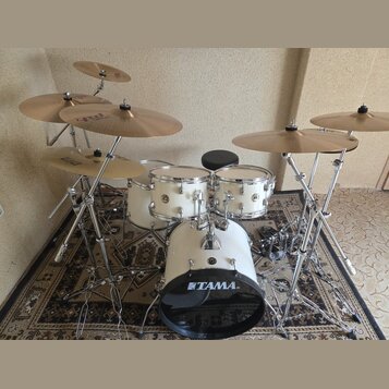 Bílá bicí souprava značky TAMA s chromovaným kováním. Sestává z basového bubnu s nápisem TAMA, dvou tomů, malého bubnu a několika činelů na chromovan…
