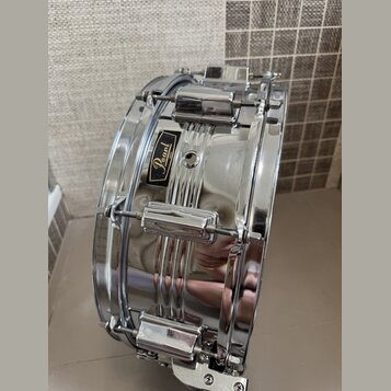 Stříbrný chromovaný malý buben s kovovým korpusem. Na boku je umístěn černý štítkový nápis Pearl JAPAN. Povrch je vysoce lesklý s viditelnými odrazy…