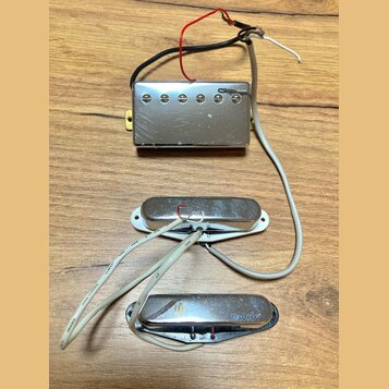 Tři elektrické snímače kytary s kovovými kryty v stříbrné barvě, umístěné na dřevěném povrchu. Horní snímač je typu humbucker s šesti viditelnými pol…