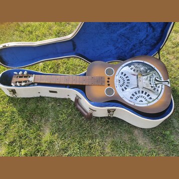 Poloklopený bílý tvrdý kufřík s modrým vnitřkem, ve kterém leží kytara typu resonator. Nástroj má hnědý přechodový povrch (sunburst), kovový kruhový…