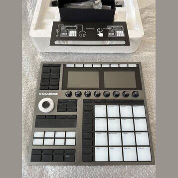 Šedý sampler Maschine+ s dvěma displeji, několika ovládacími knoflíky a bílými podsvícenými pady. Na levé straně je vidět nápis MASCHINE+ a ovládací…