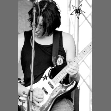 Černobílá fotografie zachycuje hudebníka s elektrickou kytarou. Kytara má tmavý lakovaný povrch, bílý pickguard a světle hnědý hmatník. Na těle kytar…