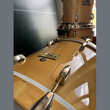 Detailní pohled na světlý, žlutohnědý lakovaný basový buben s kovovým kováním. Na korpusu je viditelný nápis Pearl Masters Custom SE. Povrch laku je…