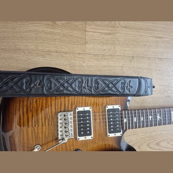 Detail elektrické kytary s vyznačenou dřevěnou strukturou a dvěma humbuckery v bílých rámečcích. Přes tělo kytary je položen černý kožený popruh s vy…