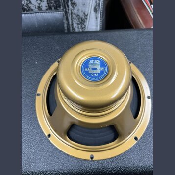 Zlatý reproduktor s kovovým rámem a středovou krytkou. Na krytce je modrý štít s nápisy CELESTION LOUDSPEAKERS, G10 ALNICO a Gold, v dolní části kruh…