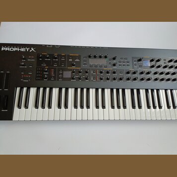 Černý syntetizátor s klaviaturou a ovládacím panelem s mnoha knoflíky, tlačítky a displeji. V levém horním rohu je bílý nápis SEQUENTIAL PROPHET X. P…