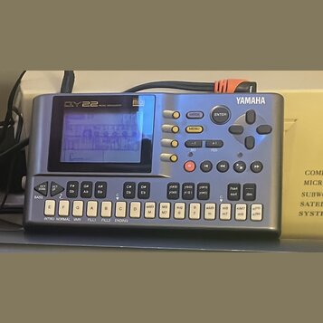 Stříbrný sekvencer s modrým displejem a ovládacími prvky. Na horní straně je nápis DY22 MUSIC SEQUENCER, logo midi a značka YAMAHA. Zařízení obsahuje…
