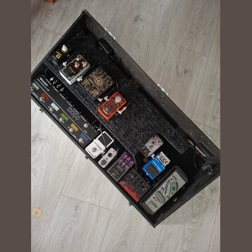 Otevřený černý transportní kufr s vnitřním pedalboardem, na kterém je umístěno několik efektů. V horní části jsou pedály s nápisy IONIK, Deluxe a v č…