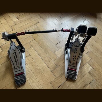 Dvojpedál na bicí v bílo-černo-červené barevné kombinaci s kovovými plátmi. Na obou plátích je černý nápis dw 9000 a pod ním text The Drummer's Choic…