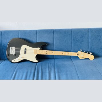 Černá elektrická basová kytara s krémovým pickguardem a světle hnědým hnedobým hmatníkem. Na hlavě je vidět nápis Squier by Fender. Nástroj má dva sn…