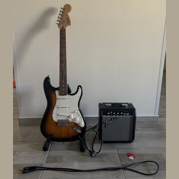 Elektrická kytara s trupem v barevnosti sunburst, bílým pickguardem a hnedým hmatníkem. Na hlavě kytary je nápis Fender. Vedle kytary stojí černý kom…