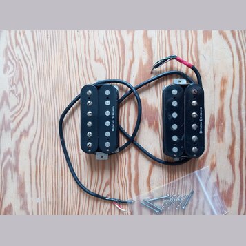 Dvě černé humbucker snímače s kovovými šroubovnicemi, připojené k sobě černým kabelem s odhalenými vodiči na koncích. Na obou snímačích je svisle nap…