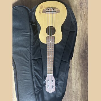 Světlý dřevěný ukulele položené na černém obalu. Na horní části korpusu je vidět hnědé logo s nápisem, který není plně čitelný. Na hlavě nástroje je…