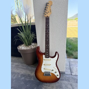 Elektrická kytara s trupem v červenohnědém sunburst provedení a bílým pickguardem. Na hlavě hmatníku je viditelné logo Fender a nápis STRATOCASTER. H…