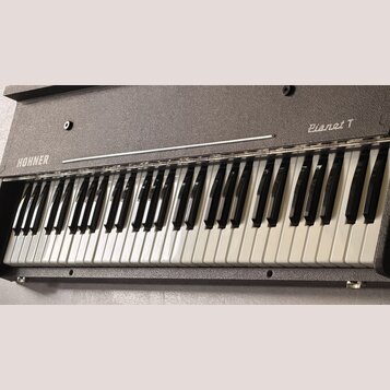 Elektrické piano s šedým, texturovaným povrchem korpusu. Na levé straně je bílý nápis HOHNER a vpravo kurzívou nápis Pianet T. Nástroj má klasickou k…