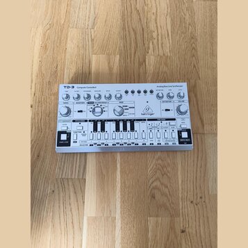 Bílá analogová basová linka syntetizátoru s nápisem TD-3 Computer Controlled Analog Bass Line Synthesizer a logem behringer. Zařízení obsahuje řadu o…