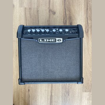 Černý kytarový kombo zesilovač značky LINE 6. Horní ovládací panel obsahuje několik otočných potenciometrů s popisky DRIVE, BASS, MID, TREBLE, CHAN V…