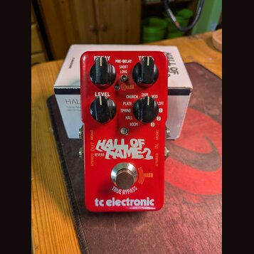 Červený efekt pedál s nápisem HALL OF FAME 2 a logem tc electronic. Na horní straně jsou čtyři černé ovládací knoflíky s popisky DECAY, TONE, LEVEL a…