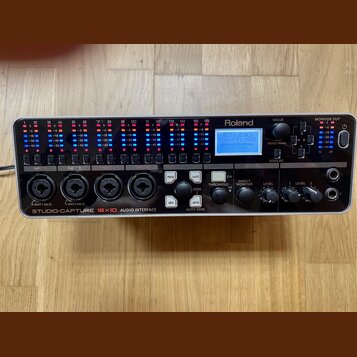 Černá racková audio rozhraní s nápisem Roland a STUDIO-CAPTURE 16x10 AUDIO INTERFACE. Zařízení má v horní části řadu barevných LED indikátorů hladiny…