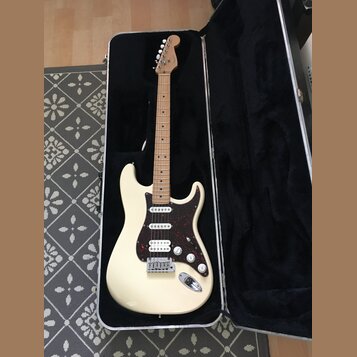 Elektrická kytara s krémovým trupem a hnědým javorovým hmatníkem v černém tvrdém kufru. Nástroj má tmavě červený pickguard, tři snímače a bílé ovláda…