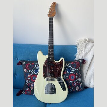 Elektrická kytara s krémovým trupem a hnědým hmatníkem. Na hlavě je nápis "by Taus". Kytara má tmavě červený pickguard s texturou želvy, tři snímače…