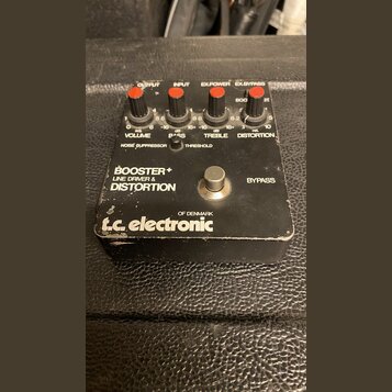 Černý efekt pedál s nápisy BOOSTER+ LINE DRIVER & DISTORTION a logem t.c. electronic OF DENMARK. Horní panel obsahuje čtyři červené oválné knoflíky s…