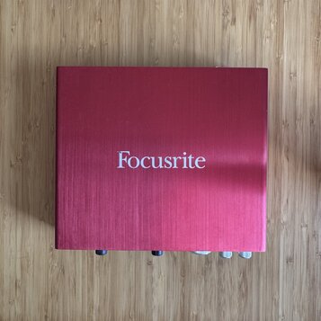 Čtvercový červený kovový box s jemně broušeným povrchem. Uprostřed horní strany je bílý nápis Focusrite. V dolní části jsou vidět části ovládacích pr…