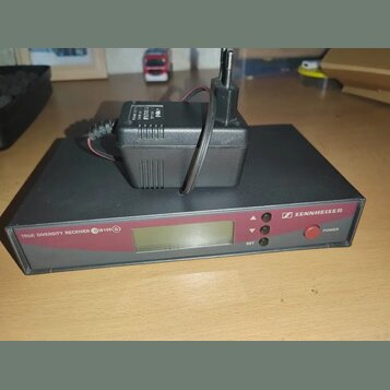 Černý bezdrátový přijímač s červeným předním panelem a LCD displejem. Na panelu jsou nápisy TRUE DIVERSITY RECEIVER EW100 G2, SET a POWER u červeného…
