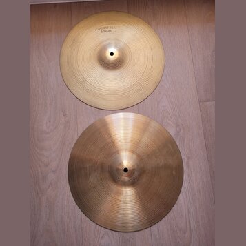 Dvě mosazné činely položené na dřevěné podlaze. Horní činel má v horní části nápis TOP NEW BEAT HI HAT. Oba činely mají jemné kruhové rýhování a vypa…