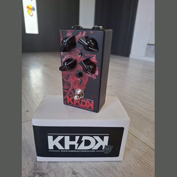 Černý kytarový efekt s červeným grafickým motivem a čtyřmi černými otočnými ovladači. Na horní straně jsou nápisy GAIN, VOLUME, BASS a TREBLE, v doln…