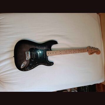 Elektrická kytara s tmavým, gradientním povrchem těla a černým pickguardem. Na hlavě hmatníku je nápis Squier STRATOCASTER. Nástroj má tři snímače, z…