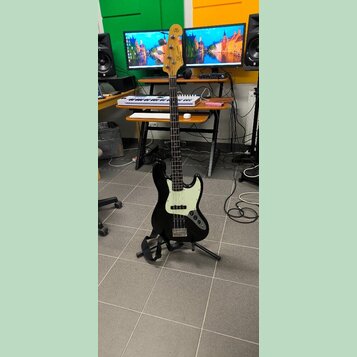 Černá elektrická basová kytara s bílým pickguardem a světle hnědým hmatníkem. Na hlavě je viditelné logo "Squier" a nápis "by Fender". Nástroj stojí…