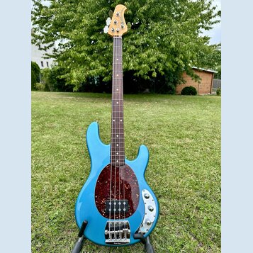 Modrá elektrická basová kytara s čtyřstrunným hmatníkem z tmavého dřeva a naturální hlavou. Tělo má v oblasti snímačů tmavý červený pickguard s textu…