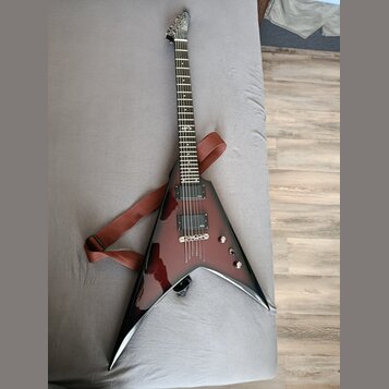 Elektrická kytara s agresivním V-tvarem v tmavě červeném průsvitném lakování. Na hmatníku je viditelné logo značky Jackson. Kytara má dva humbuckery,…