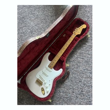 Bílá elektrická kytara typu Stratocaster s javorovým hmatníkem a zlatým hardwarem v otevřeném hnědém tvrdém kufru s červeným polstrováním. Na hlavě k…