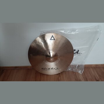 Kruhový mosazný činel s viditelným broušením povrchu. Na horní části je černý trojúhelník a nápis XIST, ve spodní části nápisy istanbul Agop Made in…