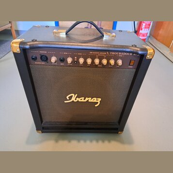 Černý akustický kytarový kombozesilovač s texturovaným povrchem, zlatými kovovými rohy a úchytem. Na předním panelu je nápis 25W ACOUSTIC AMPLIFIER T…